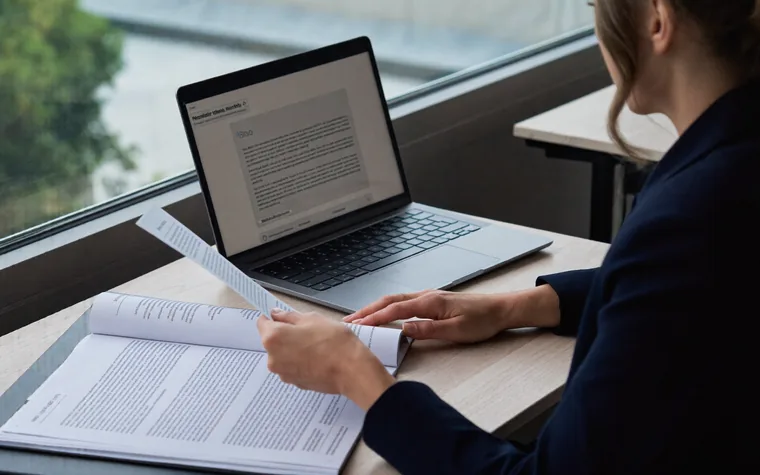 A woman at a desk reading a printed policy document beside an open laptop.